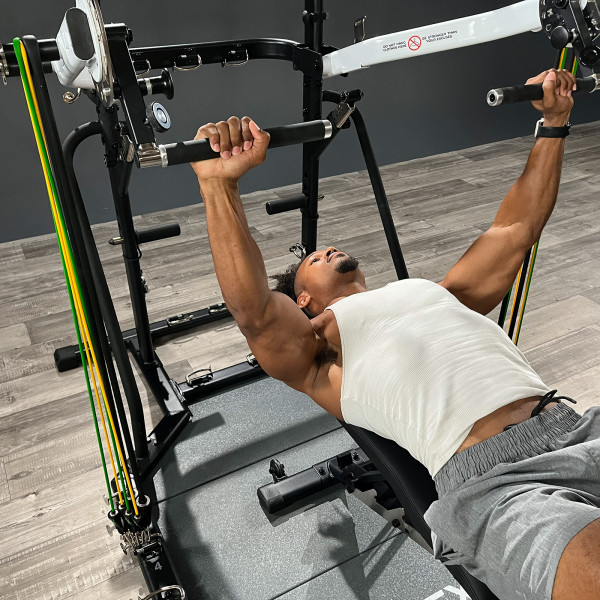 Person træner brystpres på Taurus Flexicon multimaskine med vægtstænger og elastikker i fitnessrum.